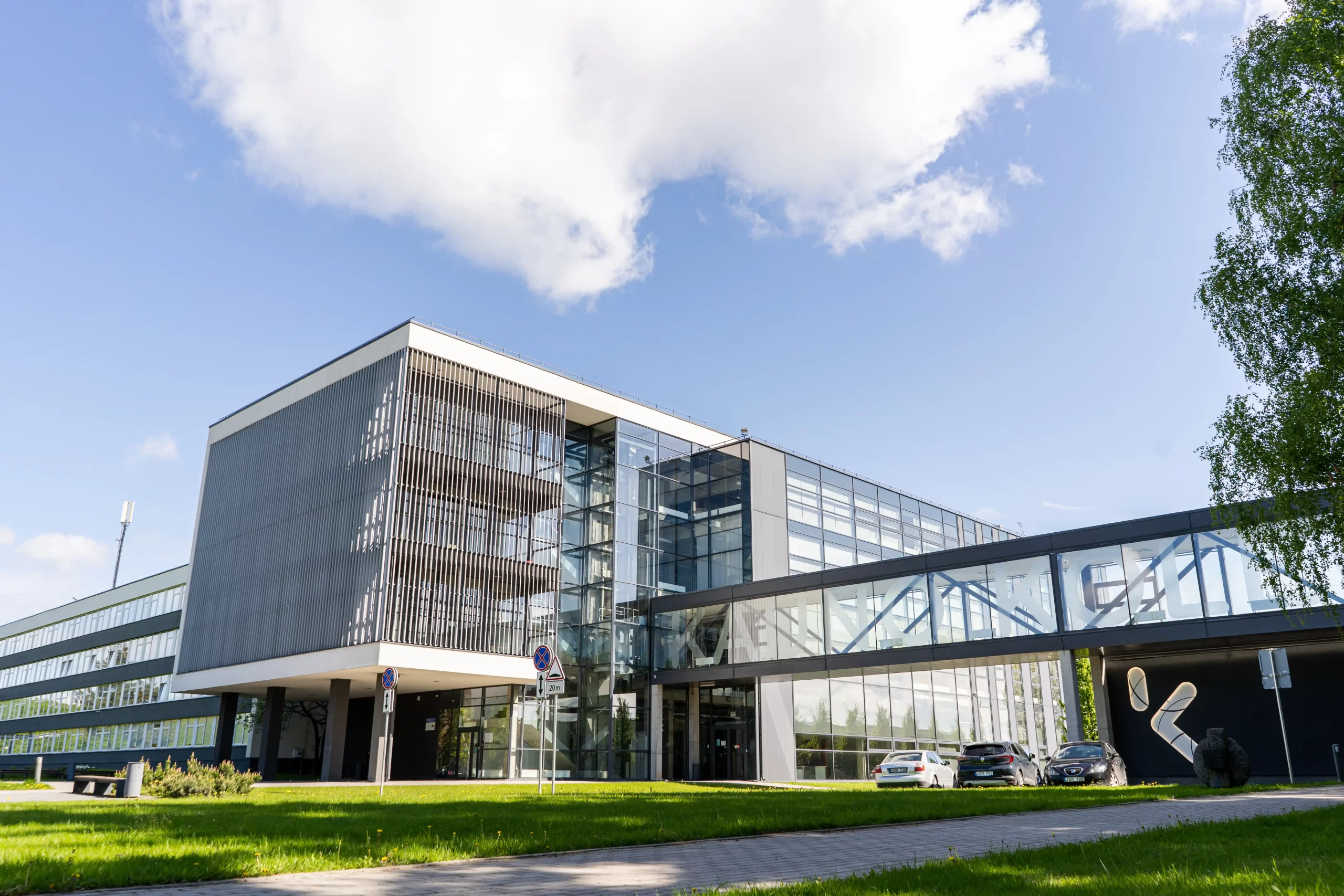 Modern exterior of the Kauno Kolegija building featuring a glass facade and a connecting skyway under a sunny sky.