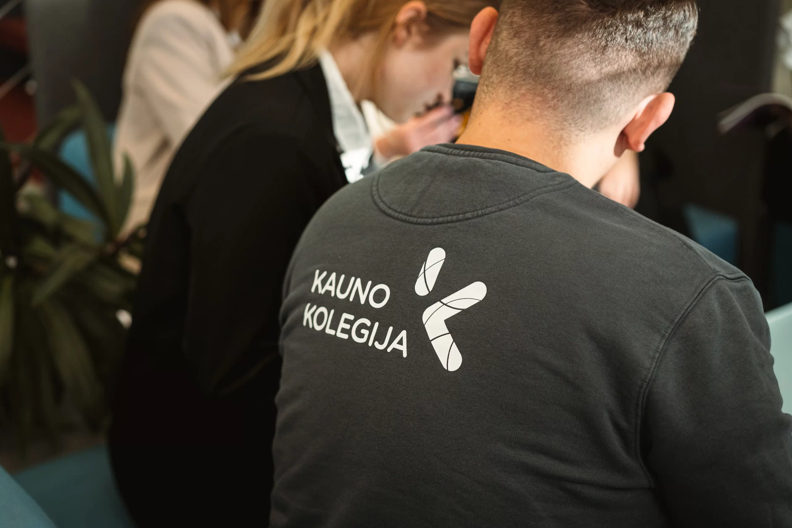 Close-up of a student's back wearing a dark grey sweatshirt with the white Kauno Kolegija logo.
