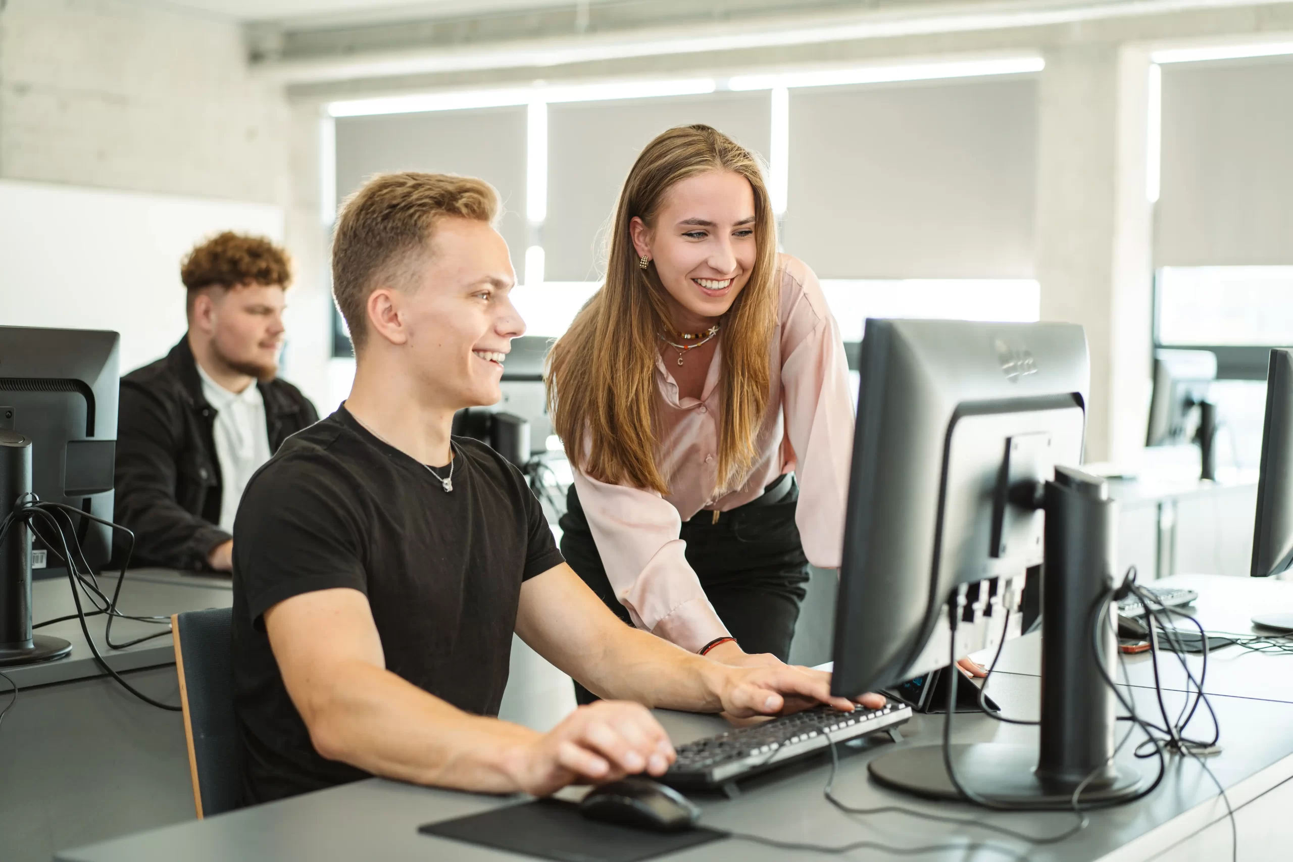 Smiling students collaborating at a desktop computer in a modern university classroom.