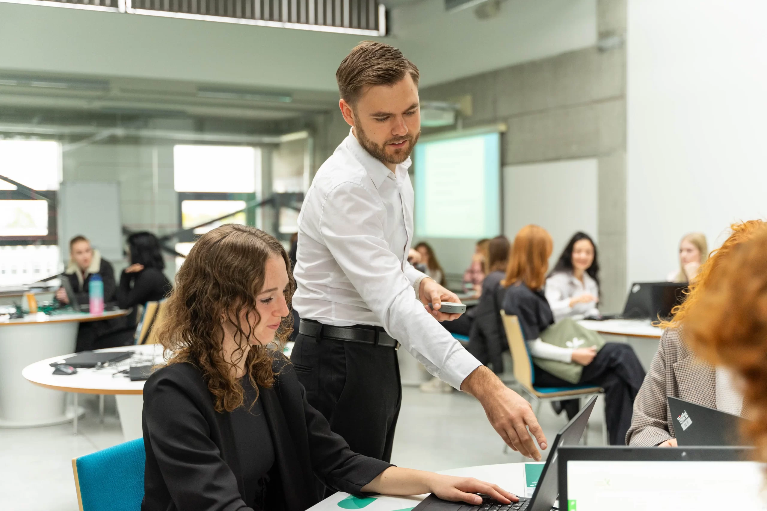 Male lecturer assisting a female student by pointing at her laptop screen in a modern classroom.