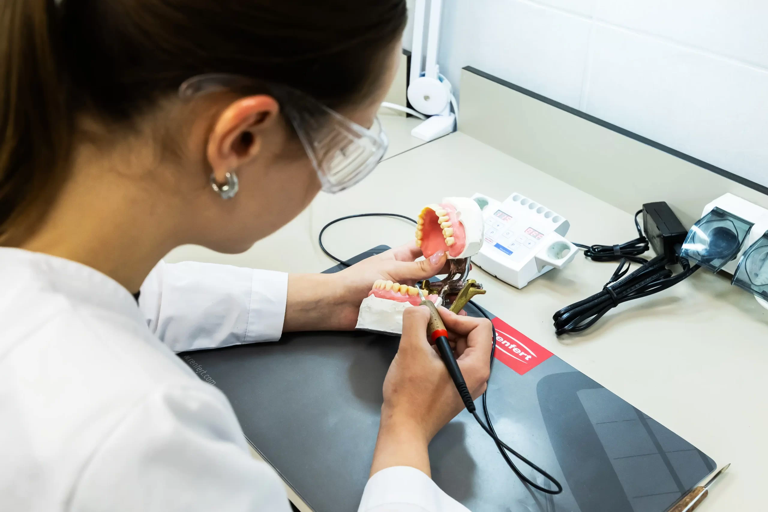 Dental technology student in safety glasses working on a denture model using a precision tool.