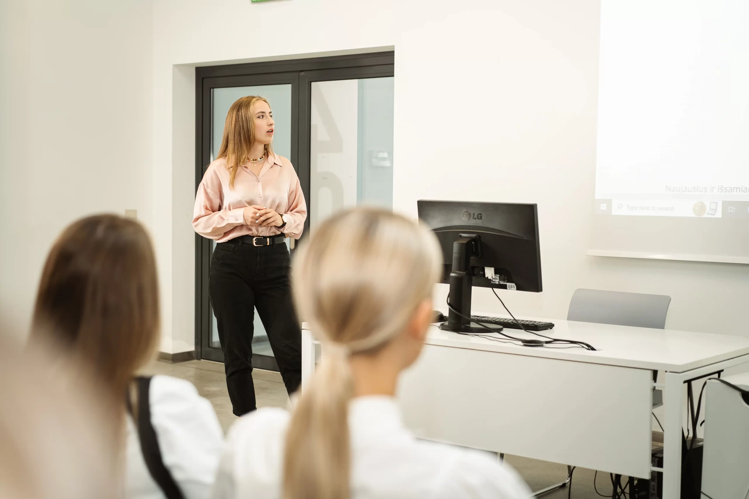 Female student giving a presentation to classmates in a modern classroom next to a projection screen.