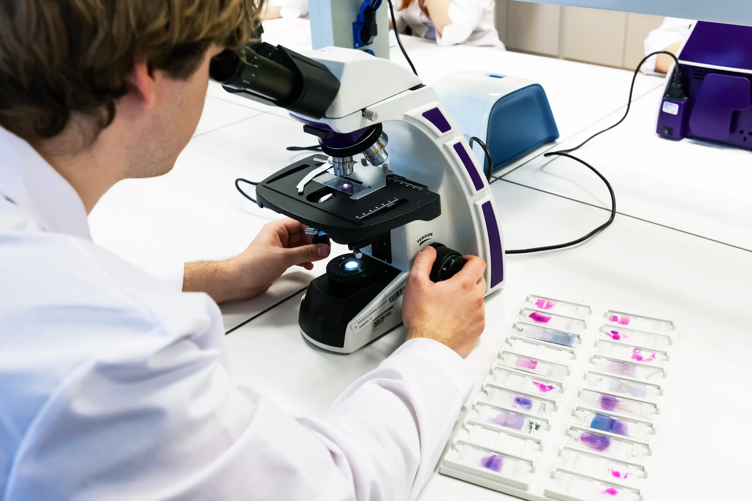 Student in a white lab coat using a microscope to examine slide samples in a science laboratory.