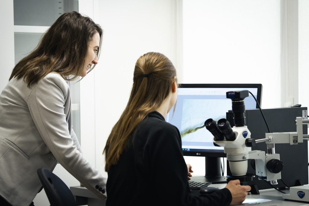 Researcher and student using a microscope in a modern laboratory at Kauno kolegija, analysing material samples on a computer screen.