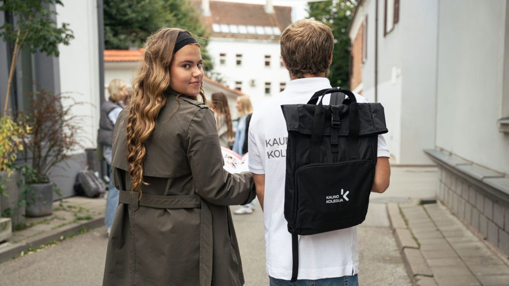 A girl with long wavy hair and a headband looks back toward the camera while walking beside a student wearing a backpack labeled ‘Kauno kolegija'.