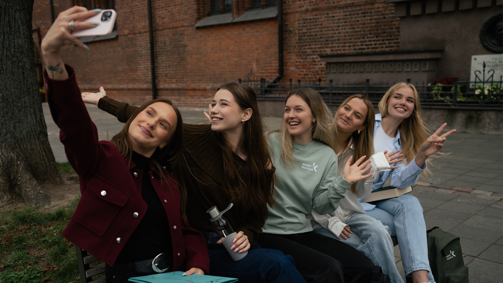 Five young women sitting on a bench outdoors in front of a brick building, smiling and posing together as one holds up a smartphone to take a selfie.