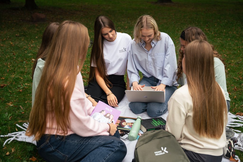 Six female students sitting in a circle on a blanket in a park, looking at a laptop together, with books, a water bottle and a backpack placed nearby.