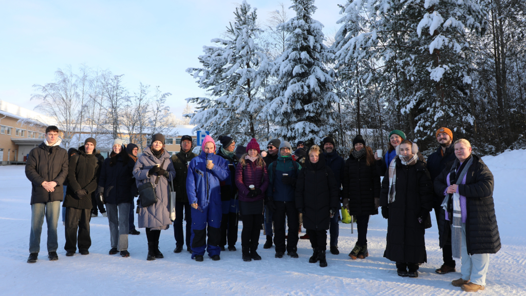 A group of people stand together outdoors on a snowy winter day. Snow-covered trees and a low building are behind them, and several people hold cups, while facing the camera in bright sunlight.