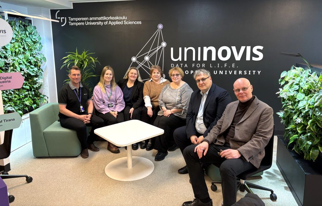 Group of representatives from Kauno kolegija and Tampere University of Applied Sciences (TAMK) seated together in a modern campus space, posing in front of a UNINOVIS wall during a partnership visit.