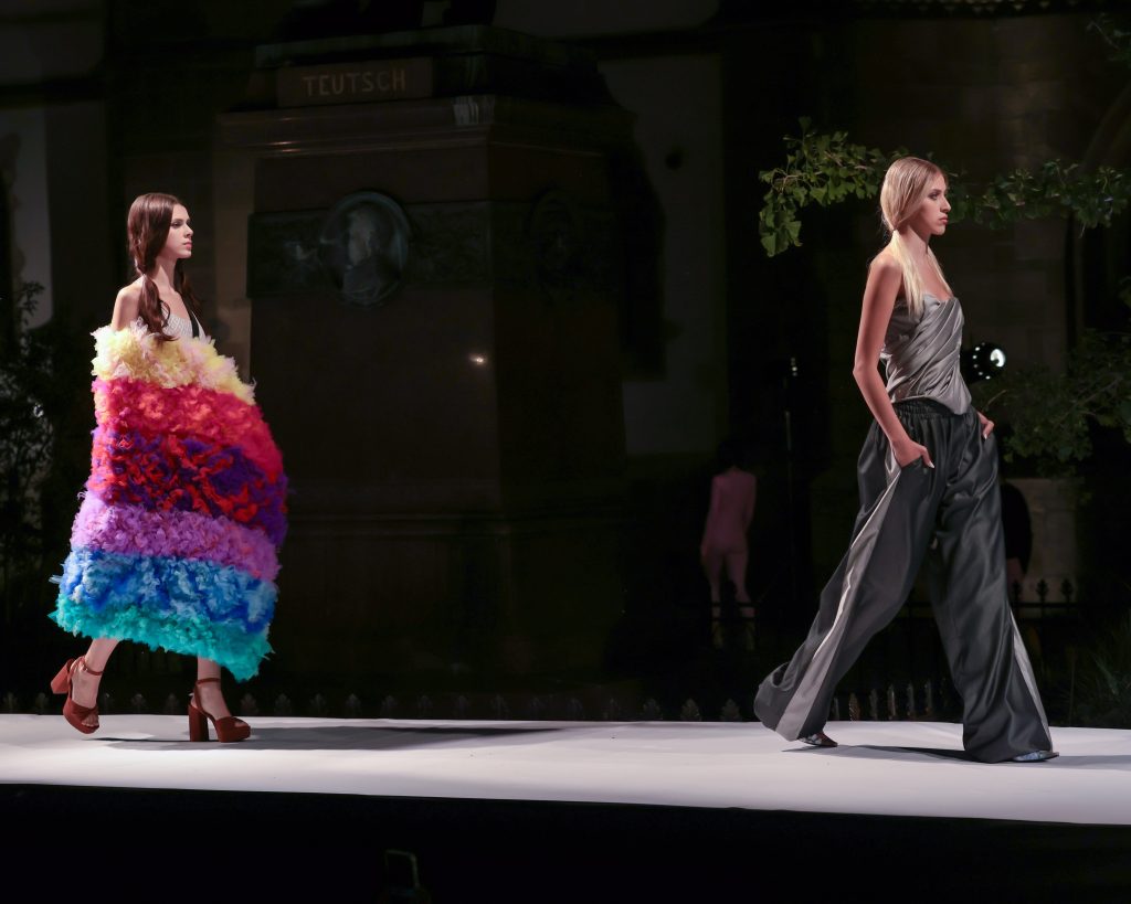 Two fashion models walk down a runway at a fashion show. One wears a dramatic, voluminous dress made of layered ruffles in bright rainbow colors with platform heels, while the other wears a sleek gray draped top with wide-leg trousers. The runway is set against a dark, elegant interior with columns and greenery in the background.