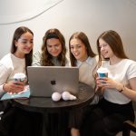 Four young women smiling and looking at a laptop screen together while sitting on a curved couch at a small round table. Two women hold coffee cups, and a black tote bag with "KAUNO KOLEGIJA" text is visible on the left.