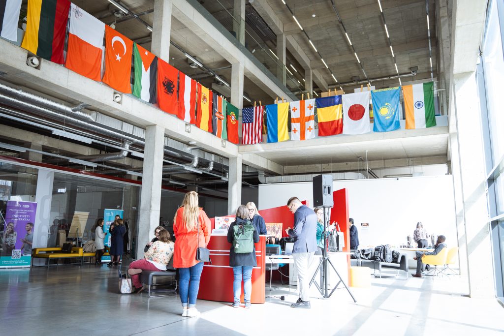 National flags of various countries hanging from a balcony in a modern hall during International Week at Kauno kolegija; participants gathered near an information desk.