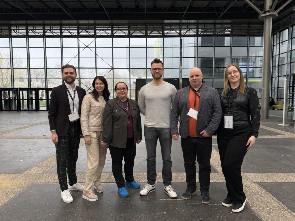 Six people stand side by side in a modern glass-walled lobby with metal structures; they are dressed in smart casual/business attire and facing the camera.