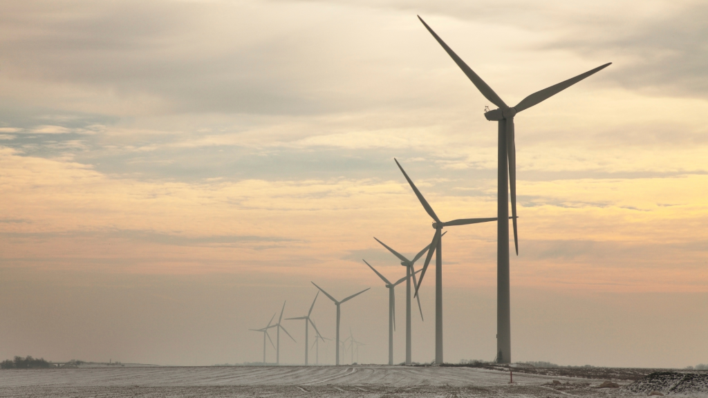 Row of large wind turbines standing in a lightly snow-covered field, stretching into the distance under a cloudy sunset sky with soft yellow and pink hues.