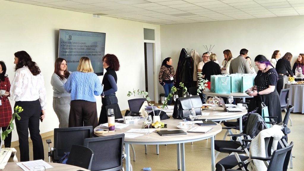 Lecturers and international participants networking in a bright classroom during a language educators’ training session, with round tables, laptops, and a presentation screen in the background.