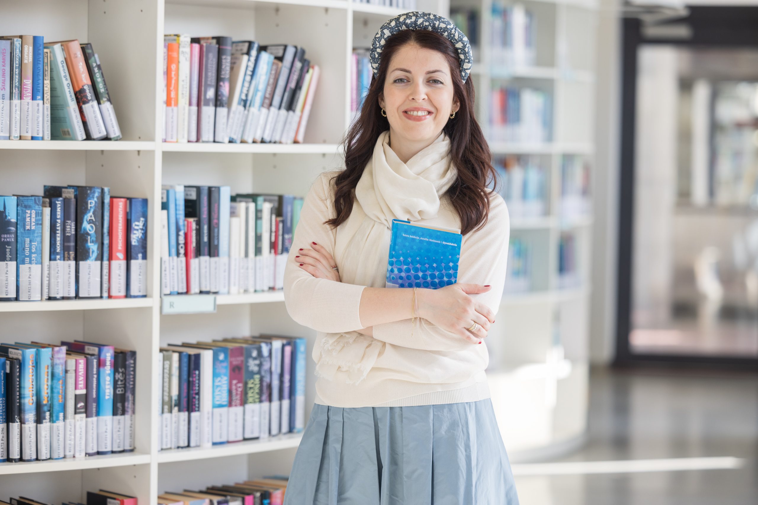 A woman stands in a library by bookshelves, smiling and holding a book in her hands.