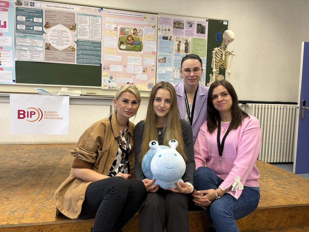 A group of four women posing together during a Blended Intensive Programme (BIP) event. They are in a classroom with academic posters and an anatomical skeleton in the background. One woman is holding a blue plushie.