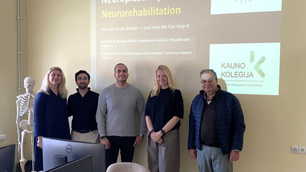 Five people standing in a classroom in front of a projected presentation slide titled “Neuroplasticity and Neurorehabilitation,” with a skeleton model and computer monitors visible in the room.