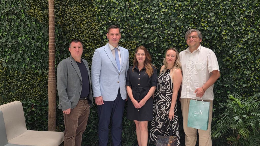 The Kauno kolegija delegation and the Chairperson of the Mexican-Lithuanian community, Jolita Mikštaitė, posing for a group photo in front of a lush green wall. A representative on the right is holding a green gift bag featuring the Kauno kolegija logo.