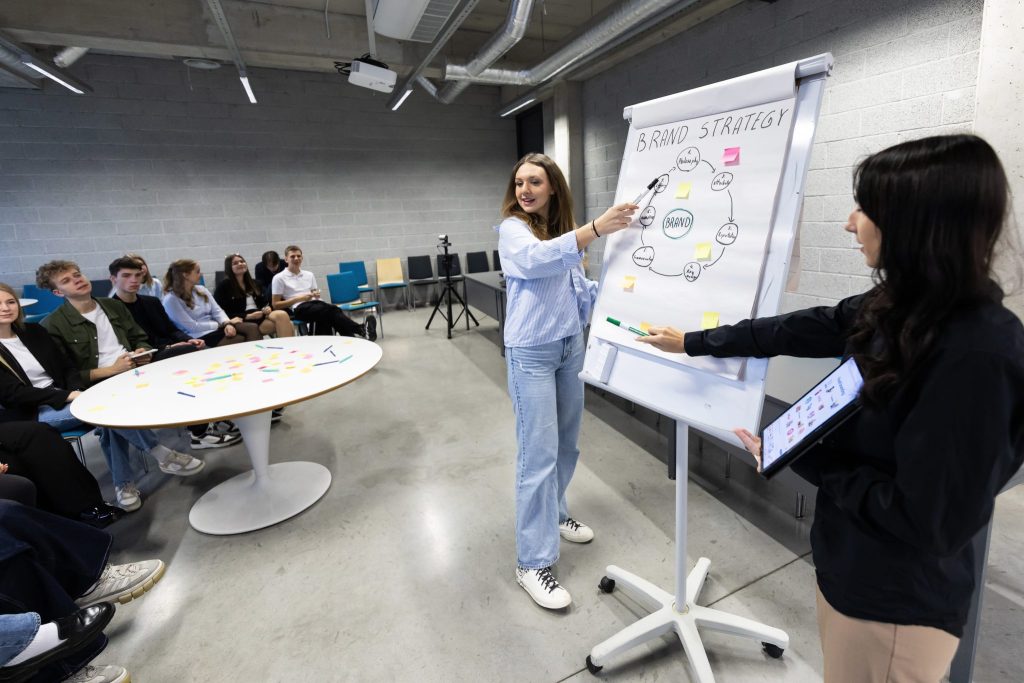 A woman presents a brand strategy diagram on a flip chart during a workshop or classroom session, while another participant assists and a group of attendees listens from seats around a table in a modern training room.