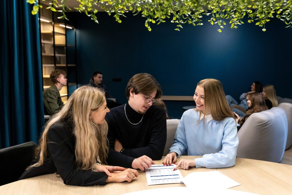 Three young adults sit around a table in a modern lounge, smiling and discussing a document together. Other people are visible in the background, and hanging greenery decorates the ceiling above.