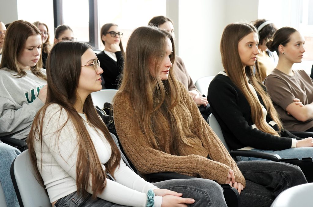 Studentės auditorijoje sėdi eilėse ir klausosi pranešimo, susitelkusios į pristatymą.
