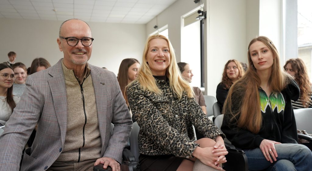 Renginio pranešėjai sėdi pirmoje eilėje ir pozuoja nuotraukai, visi žvelgia į kamerą; už jų matyti auditorijoje sėdintys studentai.