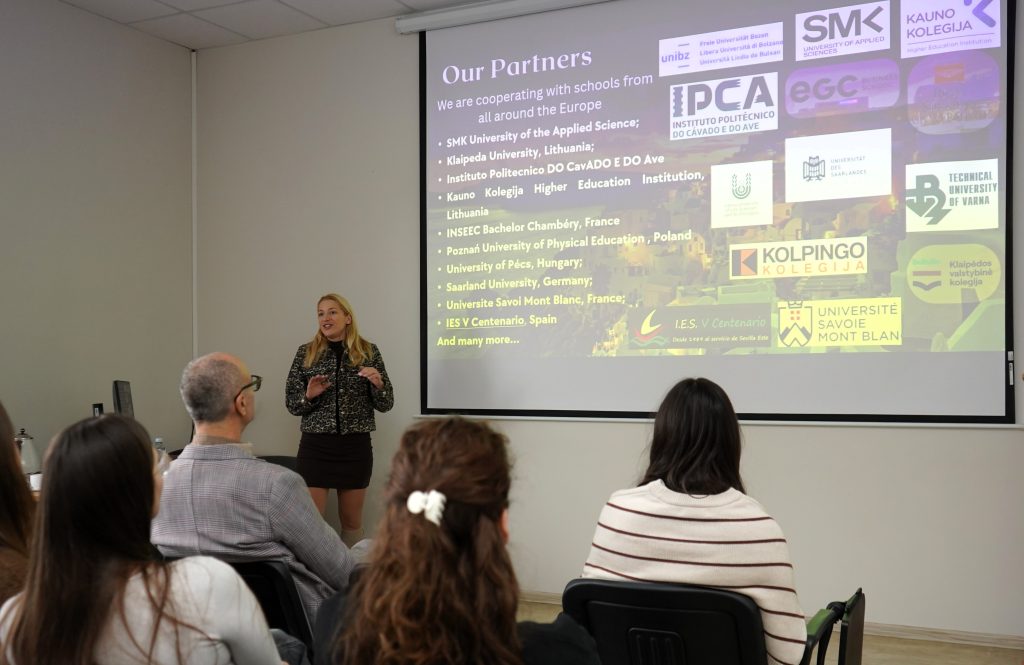 Dėstytoja stovi prie projektoriaus ekrano ir pristato skaidrę apie tarptautinius partnerius Europoje, studentai klausosi iš salės.