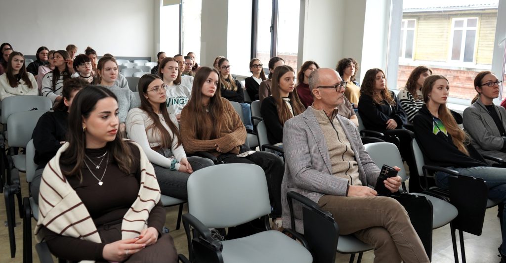 Bendras auditorijos vaizdas – studentai sėdi ir klausosi renginio, pirmose eilėse matomas užsienio pranešėjas.