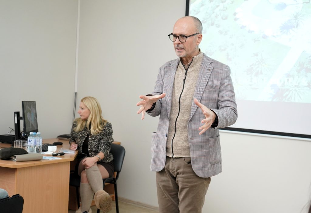 Dėstytojas stovi auditorijos priekyje ir kalba studentams, gestikuliuodamas rankomis, o už jo matomas projektoriaus ekranas