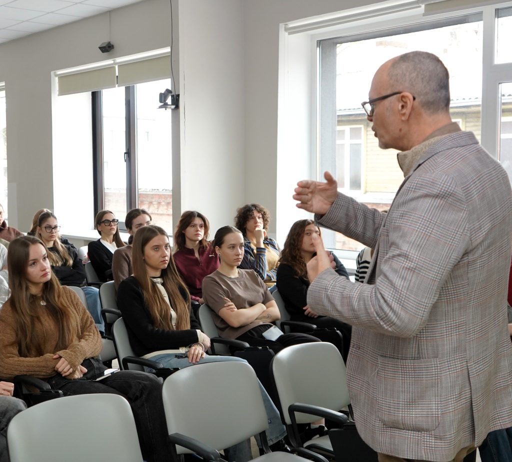 Renginio svečias auditorijoje kalba studentams, stovėdamas priekyje ir gestikuliuodamas rankomis. Studentai sėdi kėdėse ir klausosi pranešimo.