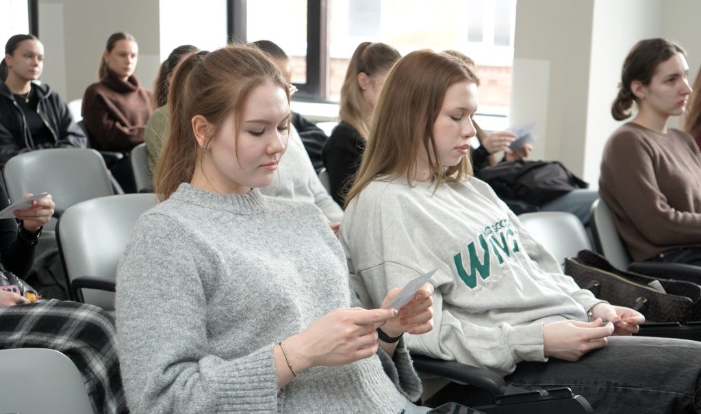 Studentės auditorijoje sėdi eilėse ir skaito rankose laikomus informacinius lapelius, susikaupusios analizuoja pateiktą medžiagą.