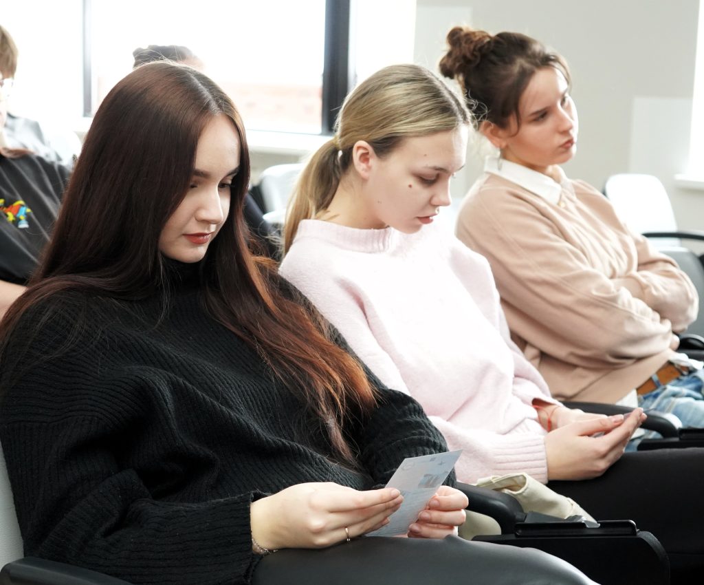 Kelios studentės pirmose eilėse skaito informacinius lapelius, sėdėdamos auditorijoje ir susitelkusios į tekstą.