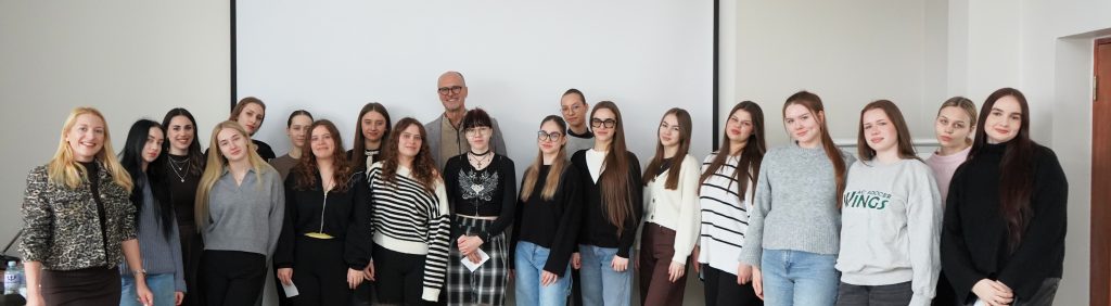 Grupinė nuotrauka auditorijoje – dėstytoja ir dėstytojas stovi kartu su studenčių grupe priešais projektoriaus ekraną. Visi pozuoja greta vieni kitų, žvelgia į kamerą, užfiksuota oficiali, bet šilta akademinės bendruomenės akimirka.