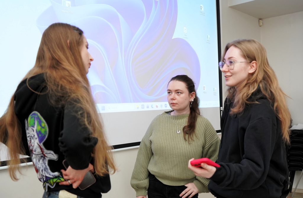 Tarptautinės veiklos koordinatorė kartu su dvejomis studentėmis, stovi prie projektoriaus ekrano ir diskutuoja tarpusavyje, viena jų rankoje laiko telefoną.