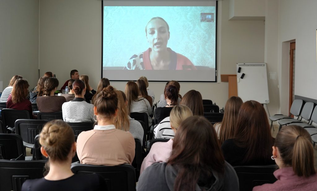 Auditorijoje studentai stebi nuotolinį pranešimą, rodomą projektoriaus ekrane, kuriame matoma kalbanti lektorė.