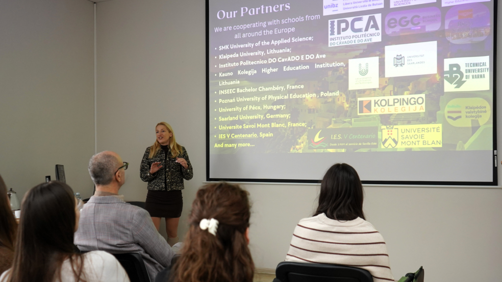 Auditorijoje vyksta pristatymas – pranešėja stovi priešais projektoriaus ekraną, kuriame rodoma skaidrė apie tarptautinius partnerius Europoje. Ji kalba susirinkusiems klausytojams, sėdintiems pirmose eilėse, o skaidrėje matomi įvairių užsienio universitetų logotipai.