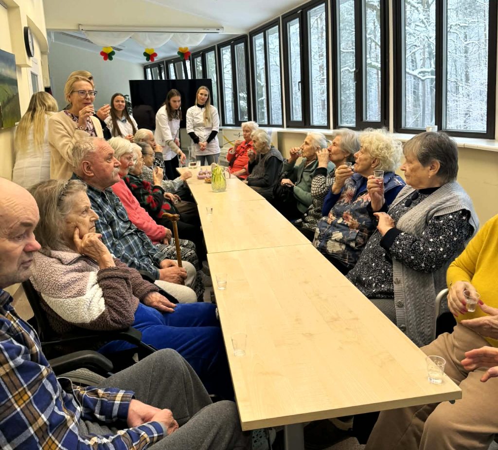 Studentės globos įstaigoje susitiko su senjorais – prie ilgo stalo susėdę vyresnio amžiaus žmonės klausosi pristatymo ir dalyvauja bendroje veikloje šviesioje patalpoje prie langų.