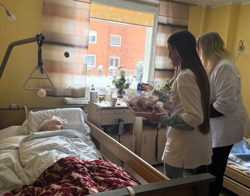 Studentės aplanko lovoje gulinčią senjorę jos kambaryje ir įteikia dovanų krepšelį. Šviesioje patalpoje prie lango vyksta šiltas susitikimas, kuriame skiriamas dėmesys bendravimui, rūpesčiui ir socialiniam palaikymui vyresnio amžiaus žmogu