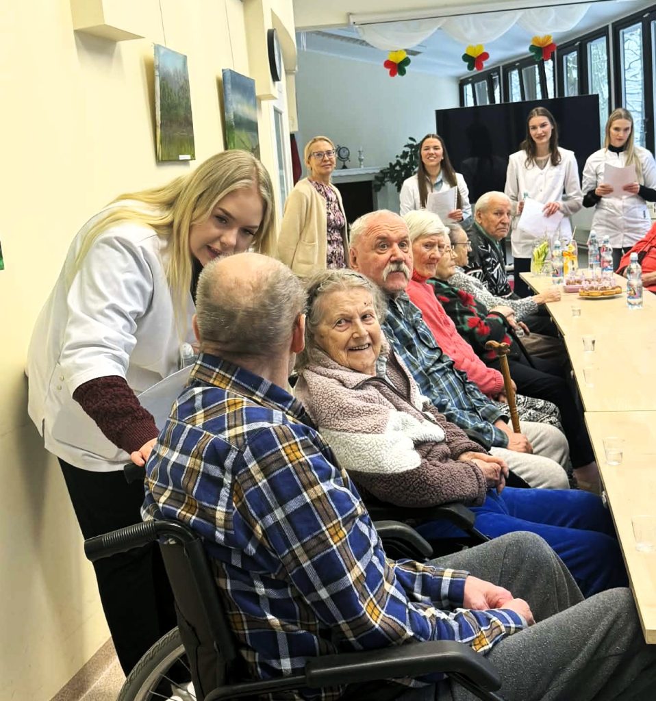 Studentė bendrauja su senjorais globos įstaigoje organizuoto susitikimo metu. Prie bendro stalo sėdintys vyresnio amžiaus žmonės dalyvauja bendruomeninėje veikloje ir klausosi studentų iniciatyvos pristatymo.