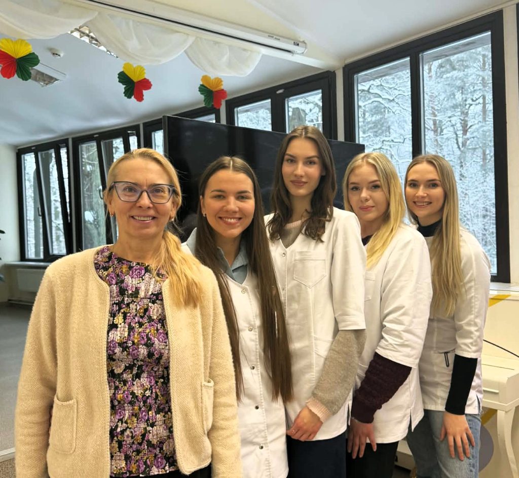 Dėstytoja ir studentės pozuoja bendroje nuotraukoje globos įstaigos patalpose. Fone matyti dideli langai ir renginio dekoracijos, o studentės vilki baltus chalatus.