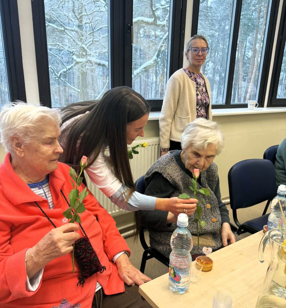 Studentė prie stalo įteikia senjorei gėlės šakelę, o šalia sėdinčios moterys stebi šią akimirką. Ant stalo matyti vandens buteliukai ir užkandžiai, susitikimas vyksta šviesioje patalpoje prie didelių langų.