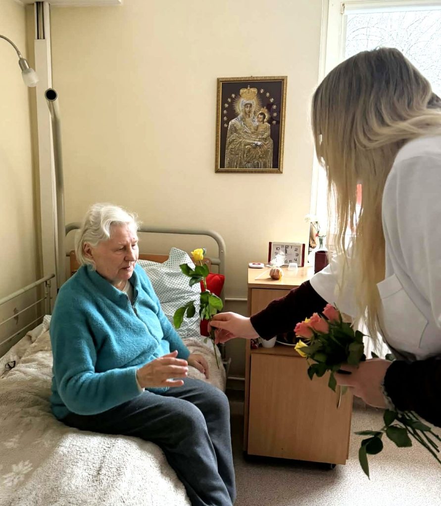 Studentė socialinės veiklos metu lanko senjorę jos kambaryje ir įteikia gėlę. Susitikimas vyksta globos įstaigoje, kur skatinamas bendravimas, dėmesys vyresnio amžiaus žmonėms ir tarpusavio ryšio stiprinimas.