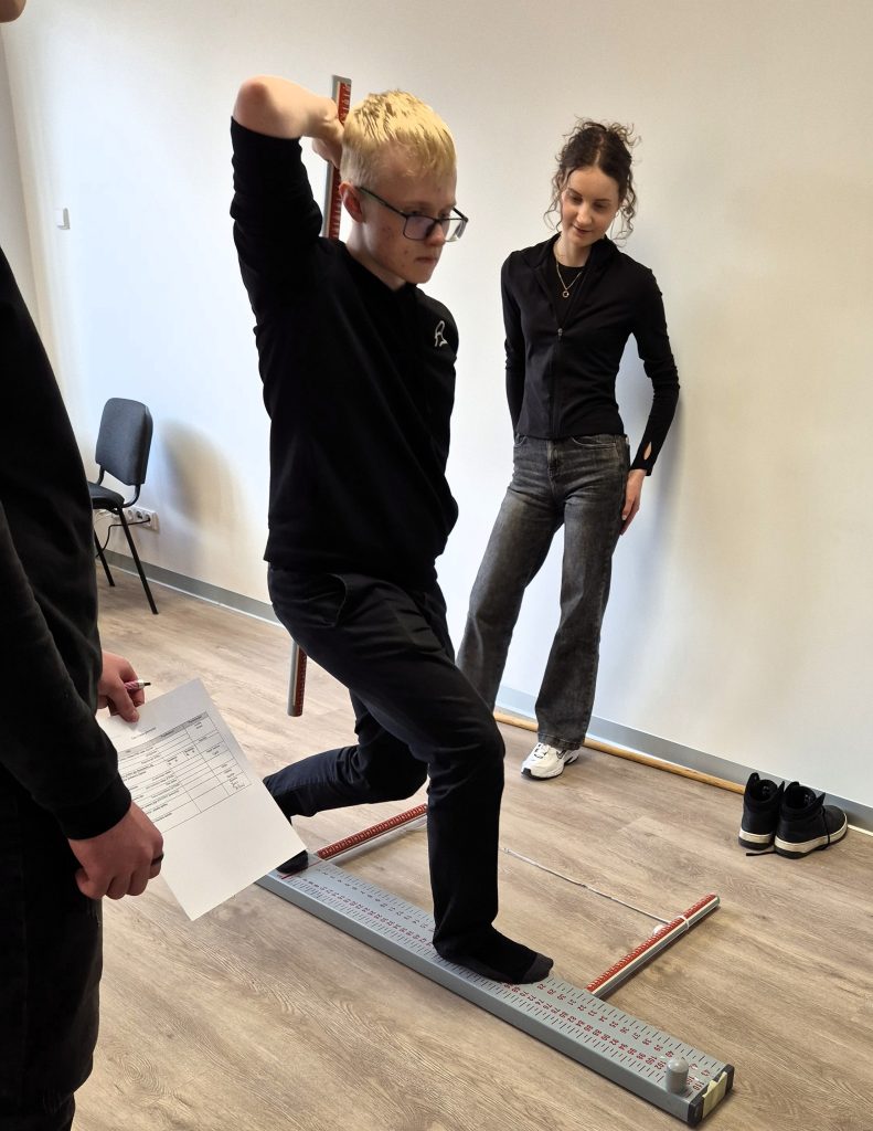 Integruotos kineziterapijos pamokos metu moksleivis atlieka pusiausvyros ir judesių kontrolės pratimą, o Kauno kolegijos studentė stebi jo atlikimą.