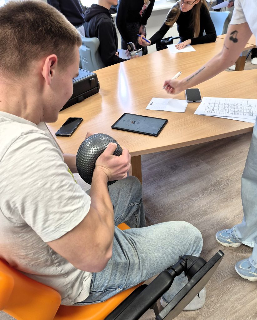 Integruotos pamokos metu moksleivis atlieka rankų jėgos ir koordinacijos užduotį su kamuoliu, o rezultatai fiksuojami šalia esančiame planešetiniame kompiuteryje.