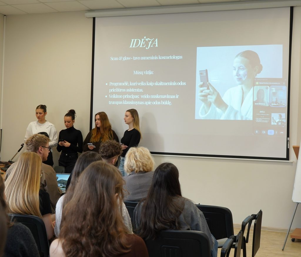 Auditorijoje keturios studentės stovi priešais ekraną ir pristato idėją konkursui, o klausytojai sėdi salėje.