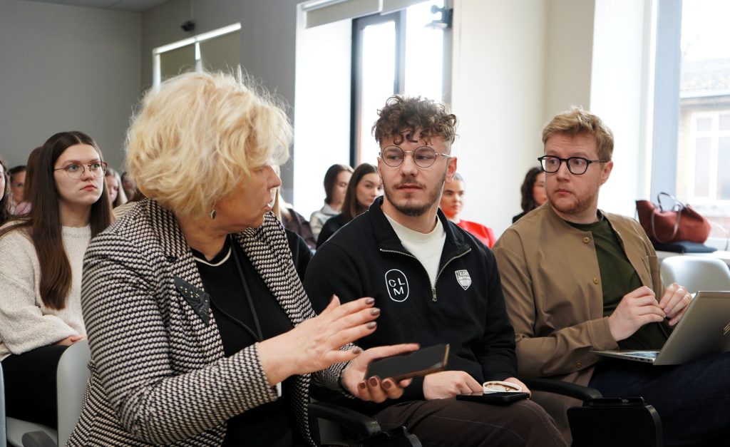 Auditorijoje sėdinti moteris kalbasi su dviem šalia sėdinčiais vyrais, o fone matyti kiti renginio dalyviai.