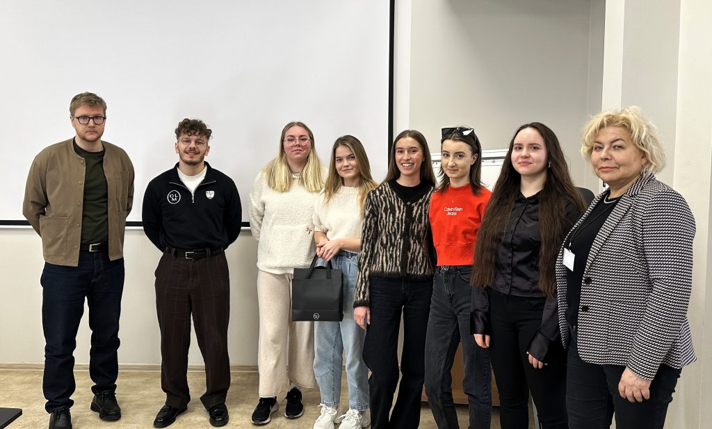 Grupė žmonių stovi auditorijoje prie ekrano ir pozuoja bendrai nuotraukai, viena mergina rankoje laiko juodą dovanų maišelį.