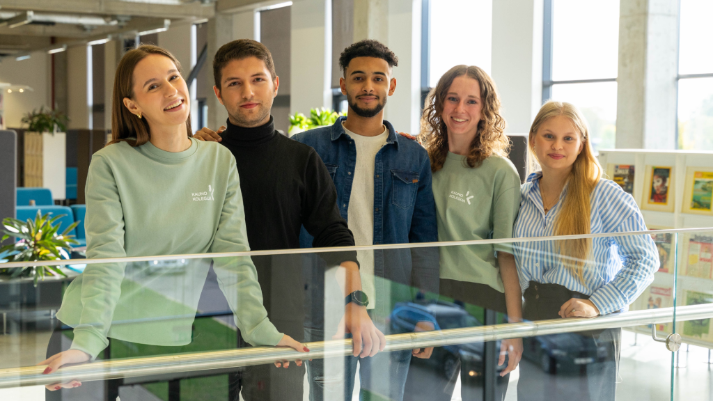 Penki Kauno kolegijos studentai pozuoja šviesioje kolegijos erdvėje prie stiklinio turėklo, vaizdas skirtas Kauno kolegijos direktoriaus vardo stipendijoms pristatyti.