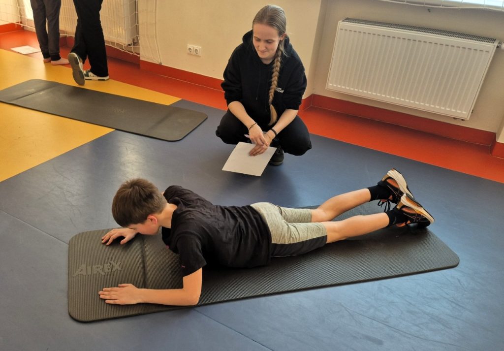 Atliekamas funkcinis stebėjimas praktinio užsiėmimo metu, kai vaikas atlieka užduotį ant kilimėlio, o Kauno kolegijos Medicinos fakulteto studentė fiksuoja rezultatus.